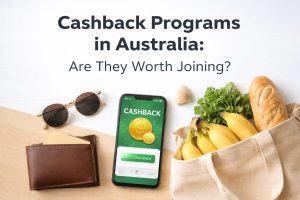 A top-down lifestyle scene showing a smartphone displaying a cashback rewards app beside a leather wallet, sunglasses, and a reusable grocery bag filled with bread, bananas, and fresh greens. The headline “Cashback Programs in Australia: Are They Worth Joining?” appears at the top of the image, illustrating how shoppers can earn rewards and savings when using a credit card for everyday purchases such as groceries and retail spending in Australia.